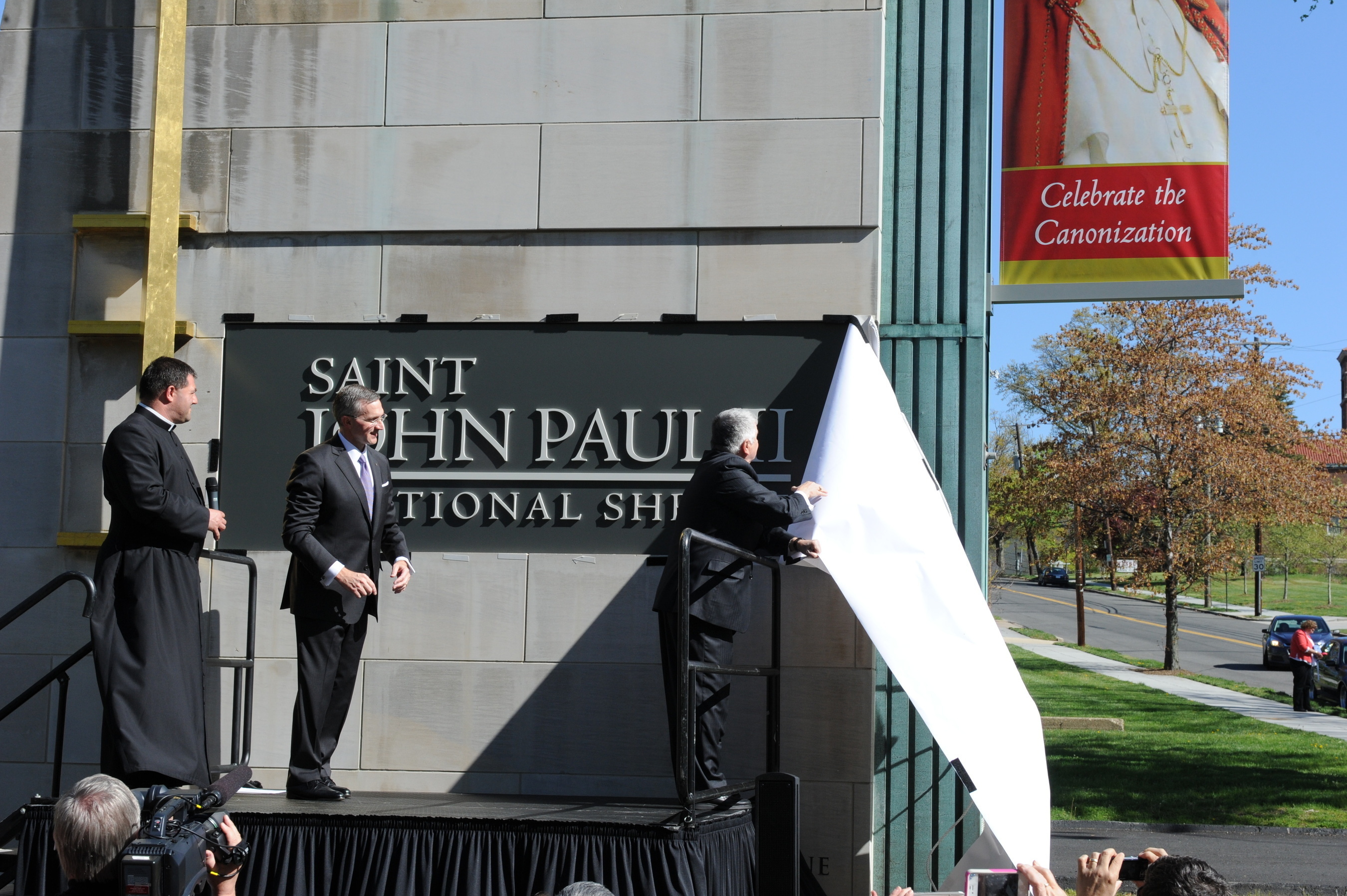 New name for the new saint.  Knights of Columbus Deputy Supreme Knight Logan Ludwig unveils a new sign for the Saint John Paul II National Shrine in Washington D.C. with the help of Shrine Director Patrick Kelly and the Rev. Gregory Gresko, shrine chaplain. Formally the shrine of Blessed John Paul II, the name upgrade followed by just a few hours the canonization of Pope John Paul, along with that of Pope John XXIII, in Rome. It is among the first places in the world to bear the name of "St John Paul II." (PRNewsFoto/Knights of Columbus)