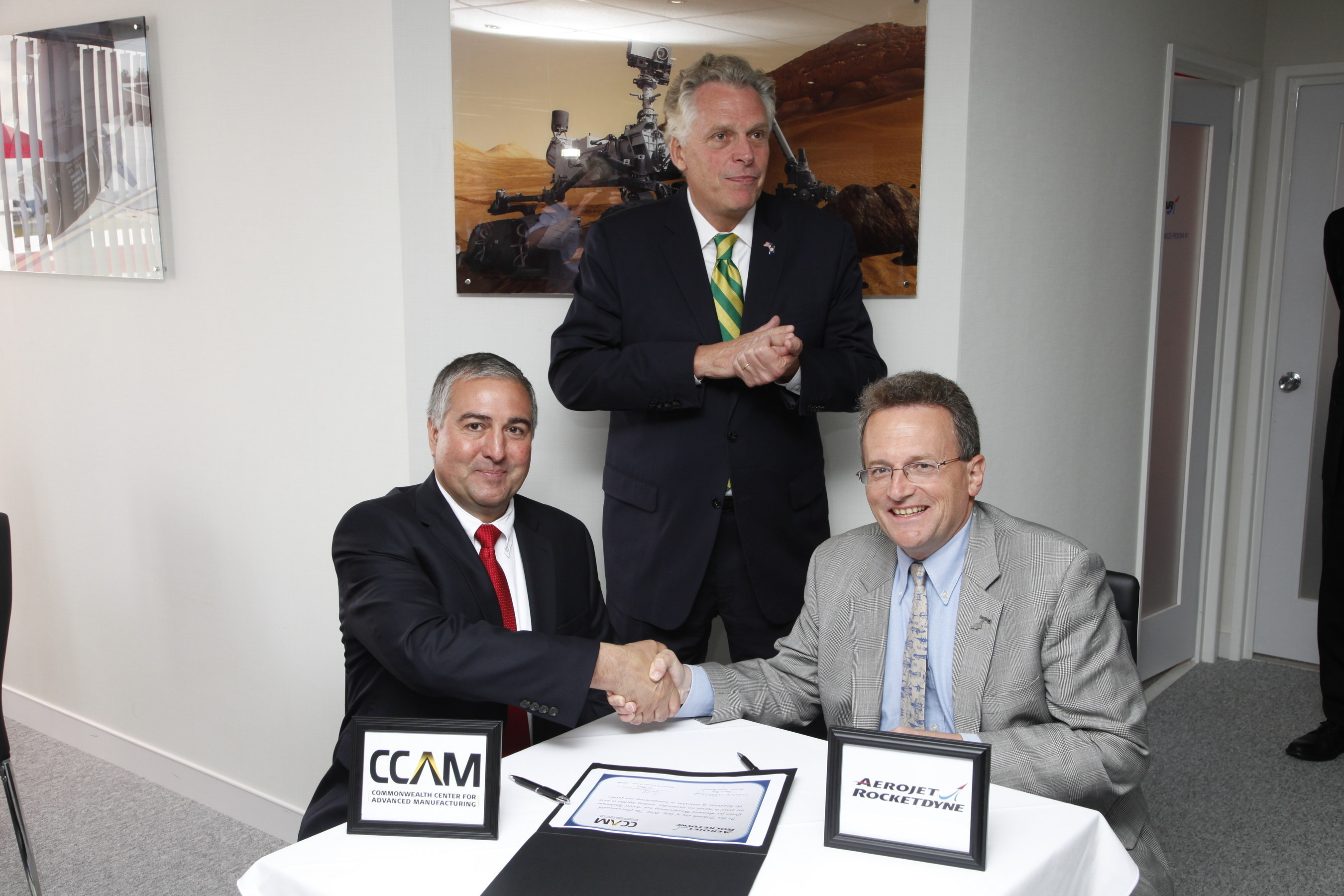 Virginia Governor Terry McAuliffe (back) oversees the agreement signed by Commonwealth Center for Advanced Manufacturing (CCAM) President & Executive Director Joseph Moody (seated, left) and Aerojet Rocketdyne President Warren Boley (right). (PRNewsFoto/CCAM)