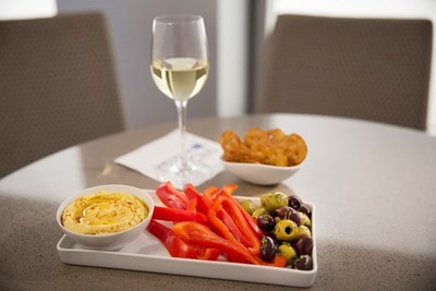 Hummus with pretzel crisps, Greek olives, freshly sliced red bell peppers, and a glass of white wine sitting on table.