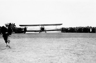 Black and white photo of airplane taking off from field, man in foreground, on April 6, 1926